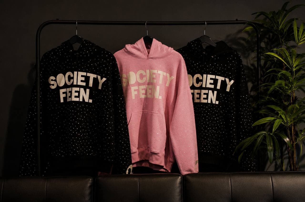 Pink and black Society Feen hoodies arranged on a clothing rack behind a dark sofa.
